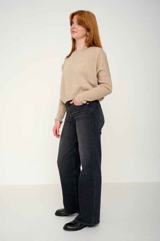 Woman wearing a oatmeal cashmere jumper with round neck and ribbed sleeves, black jeans against a plain background side view