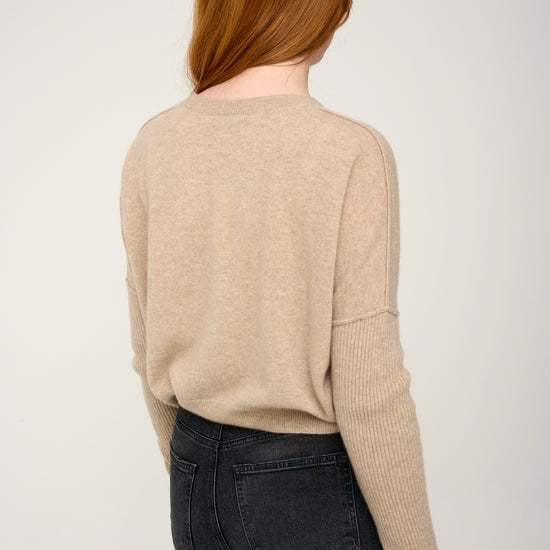 Woman wearing a oatmeal cashmere jumper with round neck and ribbed sleeves, black jeans against a plain background back view
