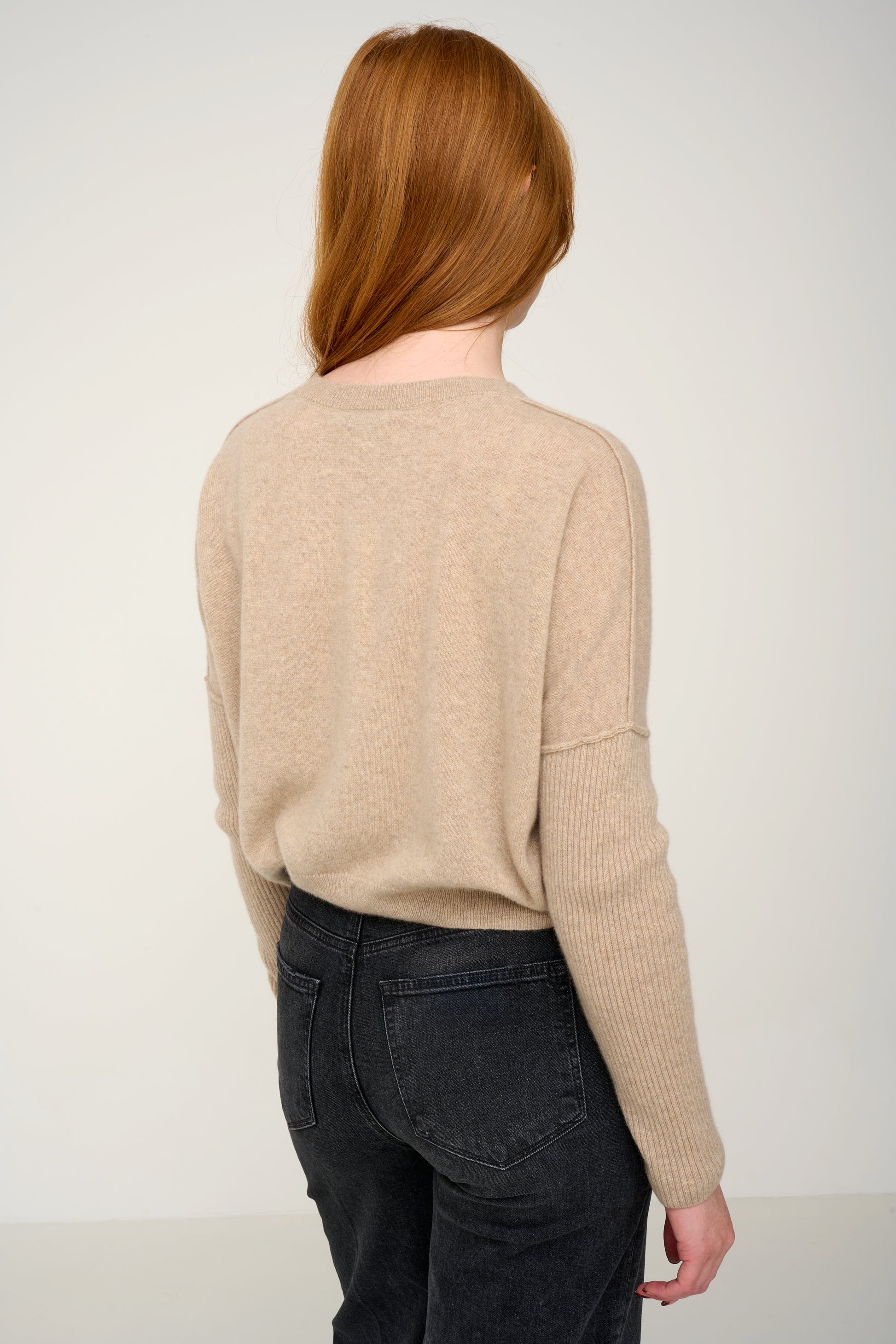 Woman wearing a oatmeal cashmere jumper with round neck and ribbed sleeves, black jeans against a plain background back view