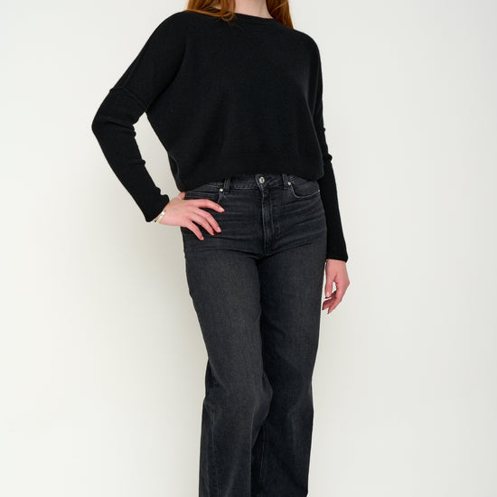 Woman wearing a black cashmere jumper with round neck and ribbed sleeves, dark jeans against a white background. Front shot.
