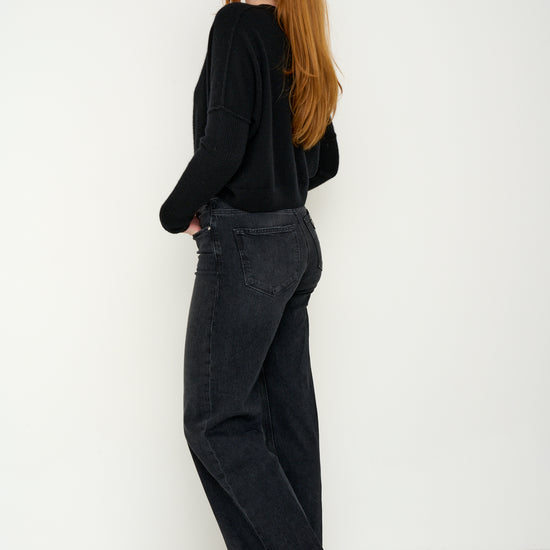 Woman wearing a black cashmere jumper with round neck and ribbed sleeves, dark jeans against a white background. Side shot.