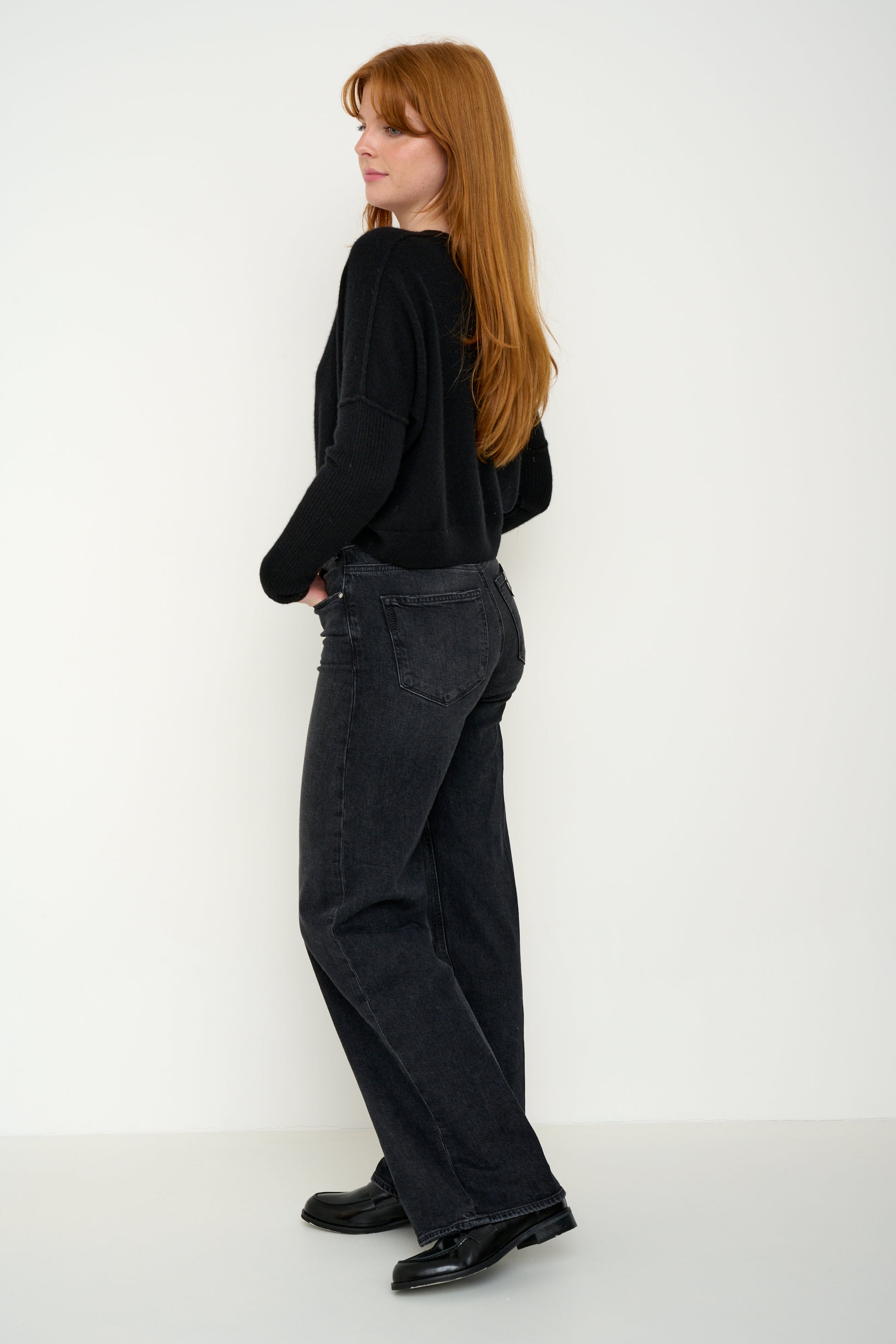 Woman wearing a black cashmere jumper with round neck and ribbed sleeves, dark jeans against a white background. Side shot.