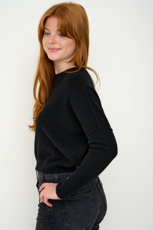 Woman wearing a black cashmere jumper with round neck and ribbed sleeves, dark jeans against a white background. Side shot.