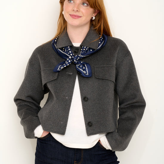 Model shot wearing grey short wool jacket with silk navy polkadot scarf layered on top in front of a white backdrop. 
