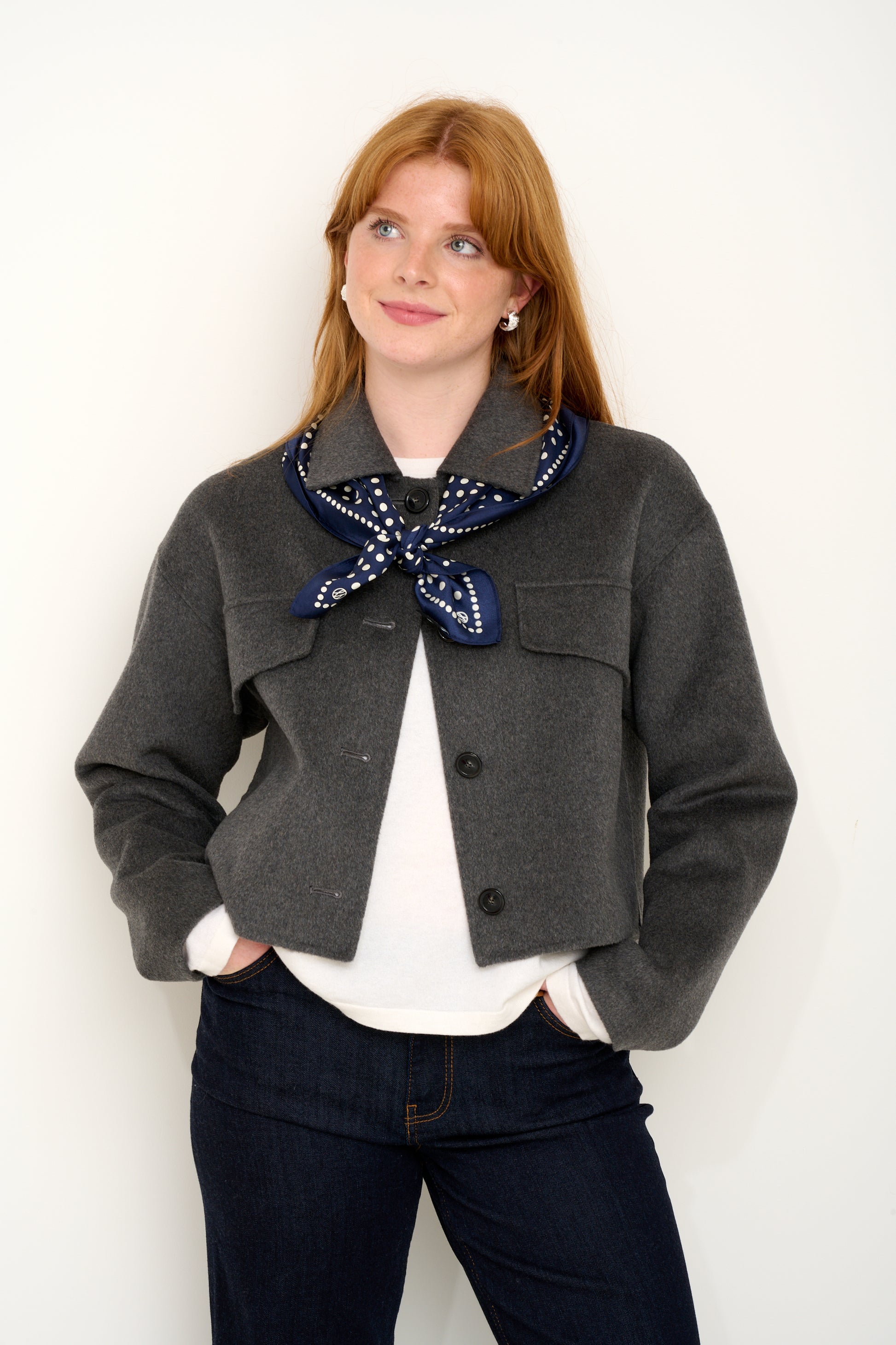 Model shot wearing grey short wool jacket with silk navy polkadot scarf layered on top in front of a white backdrop. 
