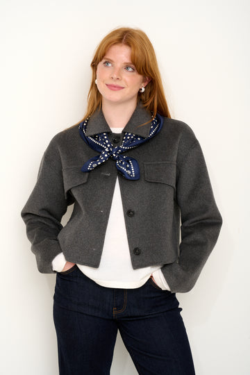 Model shot wearing grey short wool jacket with silk navy polkadot scarf layered on top in front of a white backdrop. 