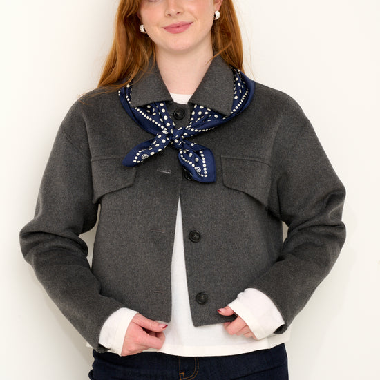 Model shot wearing grey short wool jacket with silk navy polkadot scarf layered on top in front of a white backdrop. 