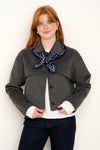 Model shot wearing grey short wool jacket with silk navy polkadot scarf layered on top in front of a white backdrop. 