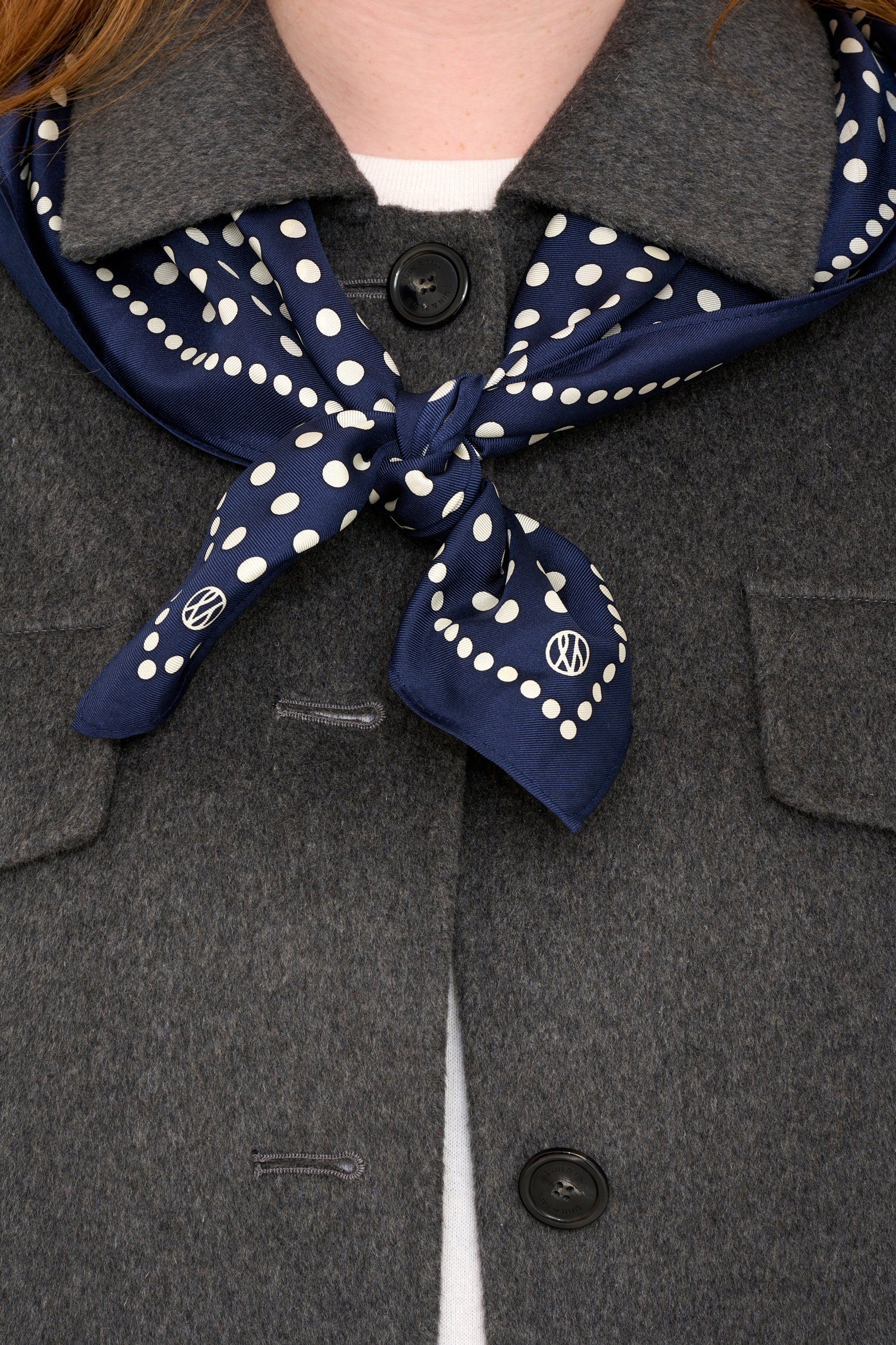 navy polka dot silk scarf tied around models shoulders layered with a grey wool jacket.