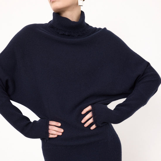 A woman wearing a dark navy blue ribbed knit turtleneck sweater with a ruffled collar detail, styled with matching ribbed knit bottoms against a plain white background.