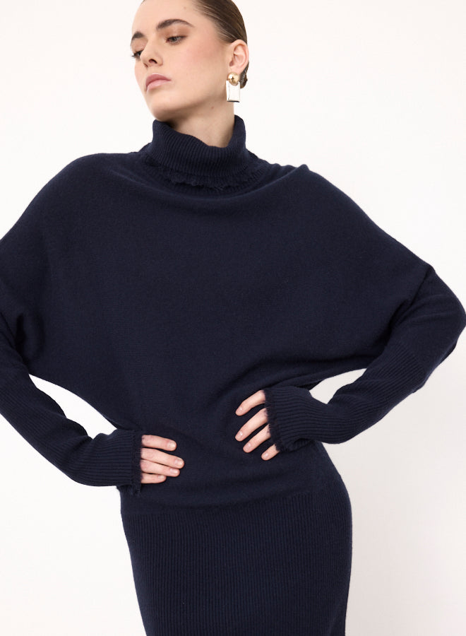 A woman wearing a dark navy blue ribbed knit turtleneck sweater with a ruffled collar detail, styled with matching ribbed knit bottoms against a plain white background.
