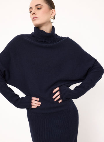 A woman wearing a dark navy blue ribbed knit turtleneck sweater with a ruffled collar detail, styled with matching ribbed knit bottoms against a plain white background.