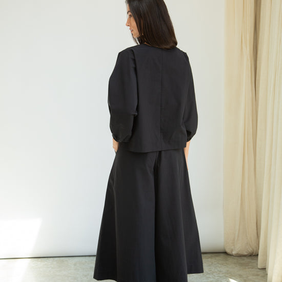 Person wearing a black outfit standing in a room with white walls and a curtain.