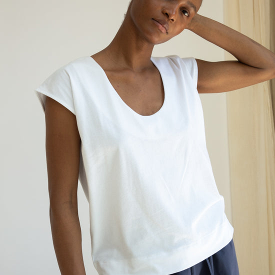 Woman wearing a white sleeveless top and dark pants against a plain background
