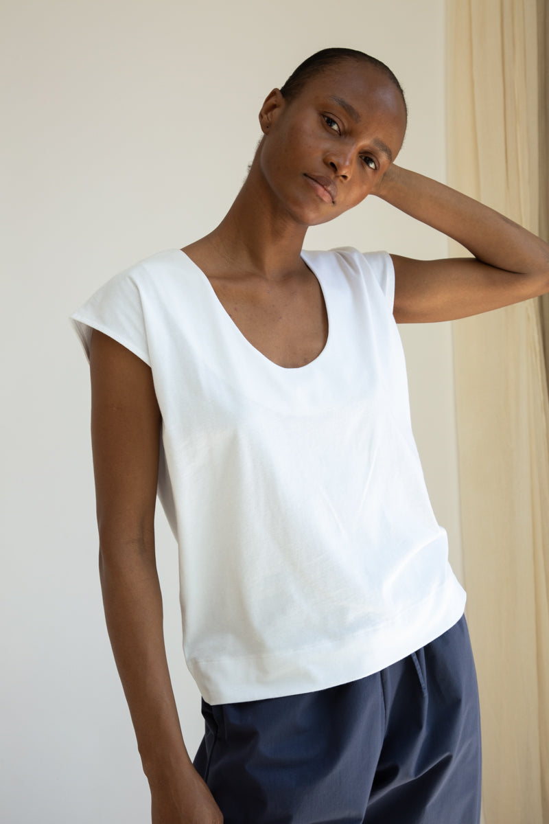 Woman wearing a white sleeveless top and dark pants against a plain background