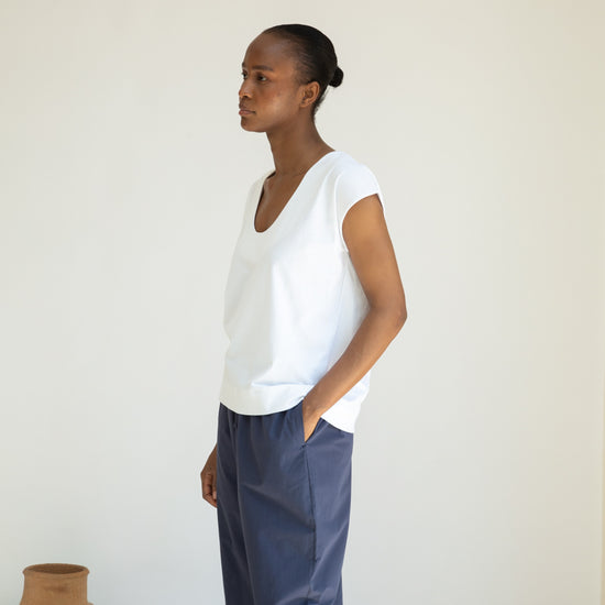 Person wearing a white top and blue pants standing against a plain wall with a vase in the foreground.