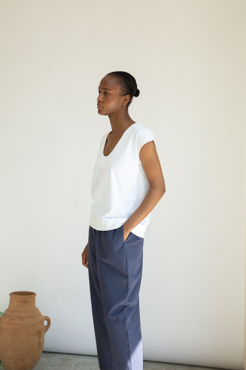 Person wearing a white top and blue pants standing against a plain wall with a vase in the foreground.
