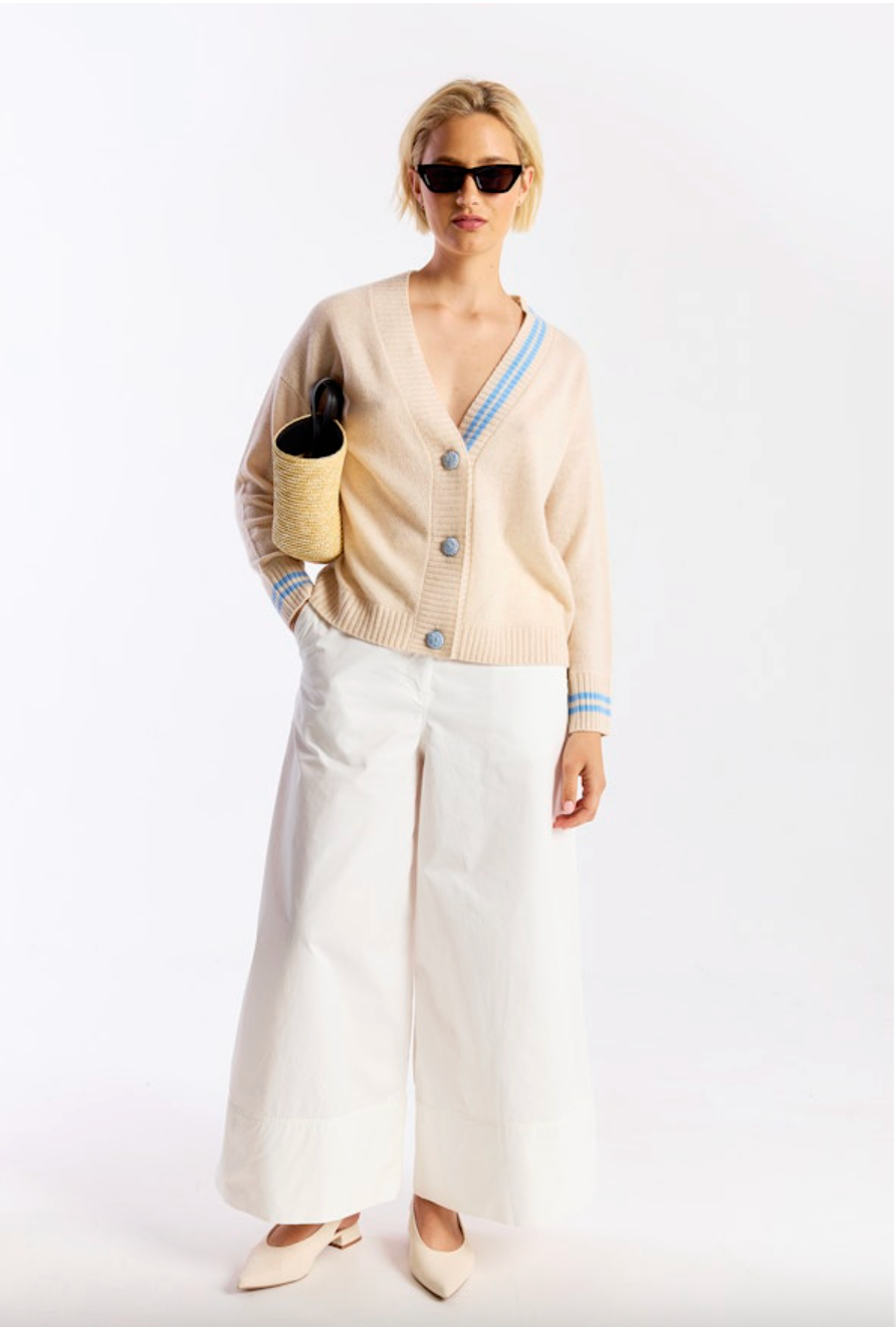Woman wearing a beige cardigan, white pants, and sunglasses on a white background