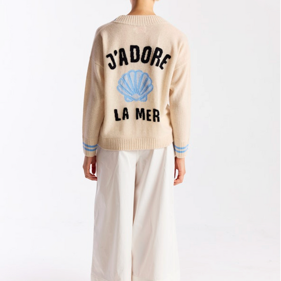 Person wearing a beige sweater with text and a shell design on the back, standing against a white background.