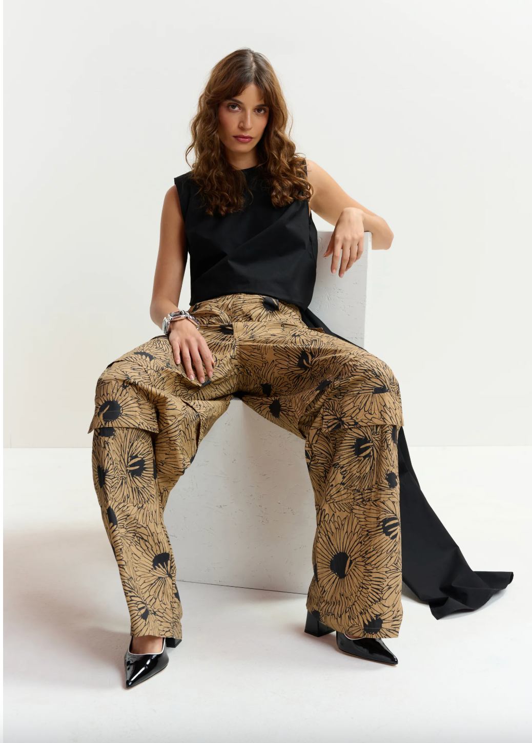 Woman sitting on a chair wearing a black top and patterned beige pants.