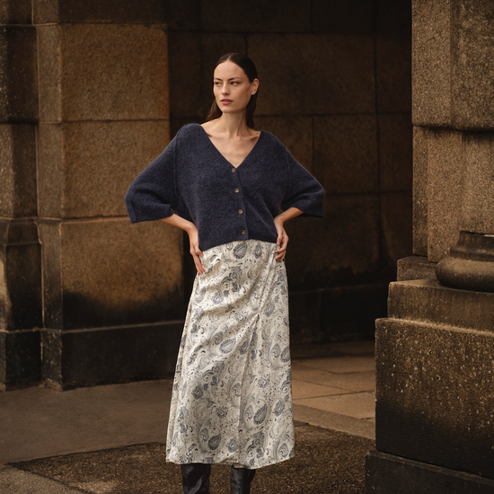Woman wearing a dark blue sweater and patterned skirt standing in front of stone columns.
