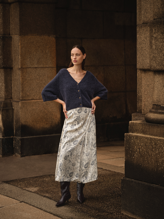 Woman wearing a dark blue sweater and patterned skirt standing in front of stone columns.