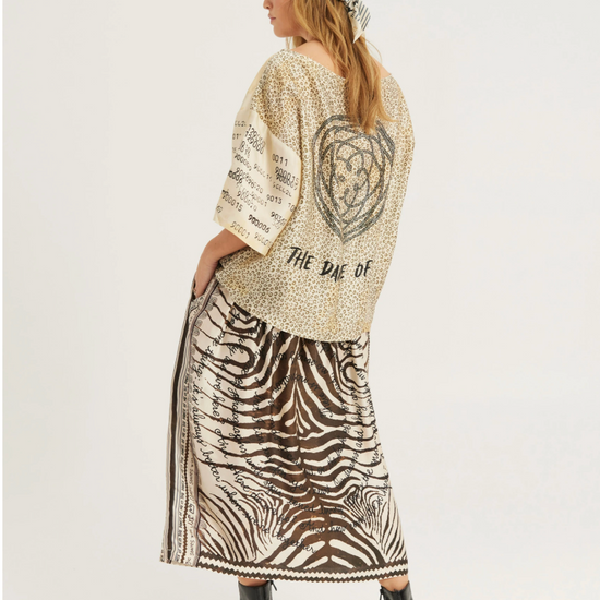 A woman shown from the back wearing a beige knit cardigan with a large black heart graphic and text, paired with a brown and white zebra-print maxi skirt and a patterned headscarf, against a plain light gray studio background.