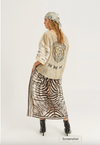 A woman shown from the back wearing a beige knit cardigan with a large black heart graphic and text, paired with a brown and white zebra-print maxi skirt and a patterned headscarf, against a plain light gray studio background.