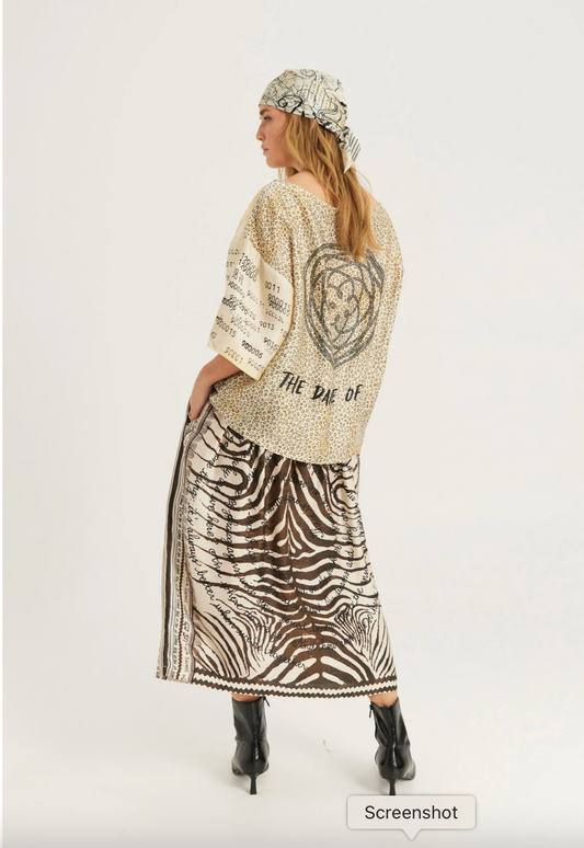 A woman shown from the back wearing a beige knit cardigan with a large black heart graphic and text, paired with a brown and white zebra-print maxi skirt and a patterned headscarf, against a plain light gray studio background.