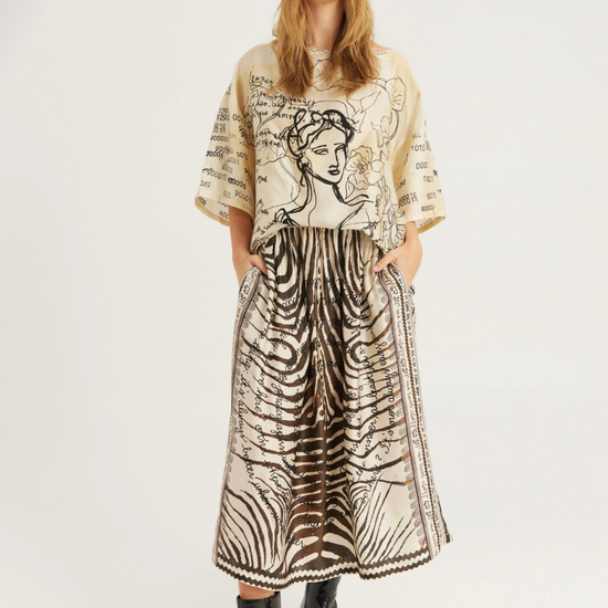 A model stands against a light gray studio background wearing a beige and black graphic print tunic top, a matching long skirt with a zebra-like pattern, a headband, and black pointed ankle boots.