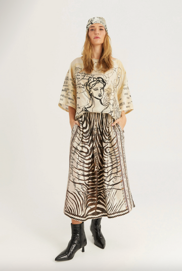 A model stands against a light gray studio background wearing a beige and black graphic print tunic top, a matching long skirt with a zebra-like pattern, a headband, and black pointed ankle boots.