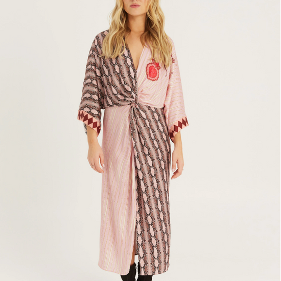 A woman modeling a long pink robe with a brown and white patterned print, featuring a red embroidered chest emblem, styled with black ankle boots against a plain white studio background.