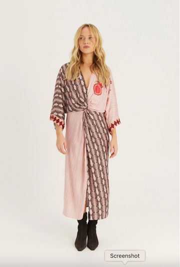 A woman modeling a long pink robe with a brown and white patterned print, featuring a red embroidered chest emblem, styled with black ankle boots against a plain white studio background.