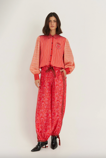 A model stands against a plain light background wearing a red and orange patterned long-sleeve top with sheer sleeves and matching red patterned wide-leg pants, paired with black pointed shoes.