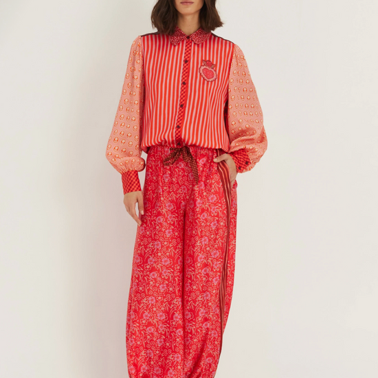A model stands against a plain light background wearing a red and orange patterned long-sleeve top with sheer sleeves and matching red patterned wide-leg pants, paired with black pointed shoes.