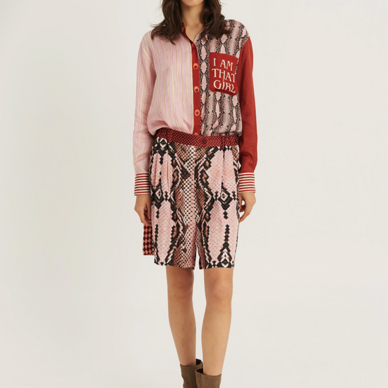 A model stands against a light gray studio background wearing a pink and red patterned button-up shirt with a snakeskin-print skirt, a red belt, and brown ankle boots. The shirt features a red patch with the text 'I AM THIS GIRL'.