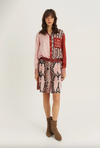 A model stands against a light gray studio background wearing a pink and red patterned button-up shirt with a snakeskin-print skirt, a red belt, and brown ankle boots. The shirt features a red patch with the text 'I AM THIS GIRL'.