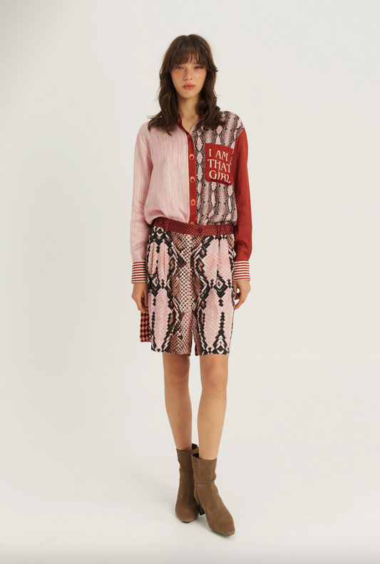 A model stands against a light gray studio background wearing a pink and red patterned button-up shirt with a snakeskin-print skirt, a red belt, and brown ankle boots. The shirt features a red patch with the text 'I AM THIS GIRL'.