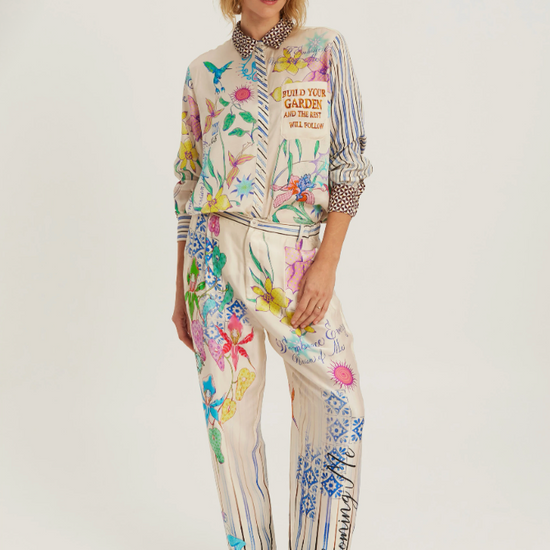 A model wearing a matching two-piece jumpsuit set with a cream base and a vibrant multicolor floral and botanical print, styled with brown heeled ankle boots against a plain light gray studio background.