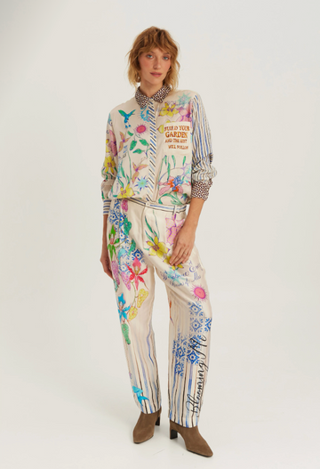 A model wearing a matching two-piece jumpsuit set with a cream base and a vibrant multicolor floral and botanical print, styled with brown heeled ankle boots against a plain light gray studio background.