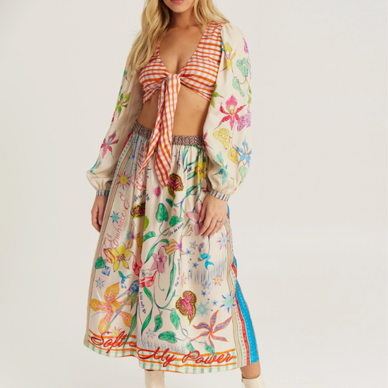 A model wearing a matching two-piece outfit featuring a red and white gingham tie-front crop top and a long-sleeved midi skirt with a colorful botanical print, styled with cream ankle boots against a light gray studio background.