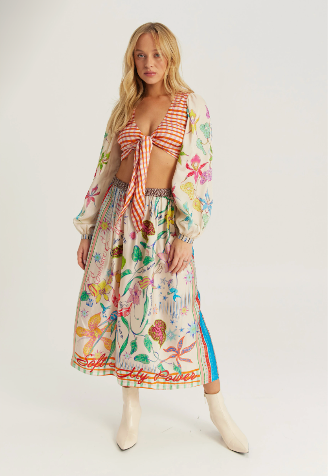 A model wearing a matching two-piece outfit featuring a red and white gingham tie-front crop top and a long-sleeved midi skirt with a colorful botanical print, styled with cream ankle boots against a light gray studio background.