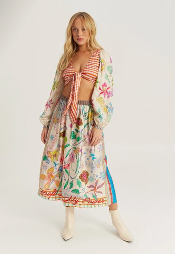 A model wearing a matching two-piece outfit featuring a red and white gingham tie-front crop top and a long-sleeved midi skirt with a colorful botanical print, styled with cream ankle boots against a light gray studio background.