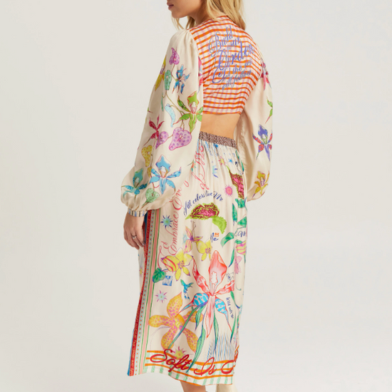 A woman modeling a long-sleeved, cropped maxi dress with a colorful floral and butterfly print on a cream base, paired with a striped crop top and white ankle boots, shown from a side-back angle against a light gray studio background.