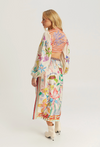 A woman modeling a long-sleeved, cropped maxi dress with a colorful floral and butterfly print on a cream base, paired with a striped crop top and white ankle boots, shown from a side-back angle against a light gray studio background.