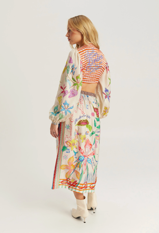 A woman modeling a long-sleeved, cropped maxi dress with a colorful floral and butterfly print on a cream base, paired with a striped crop top and white ankle boots, shown from a side-back angle against a light gray studio background.