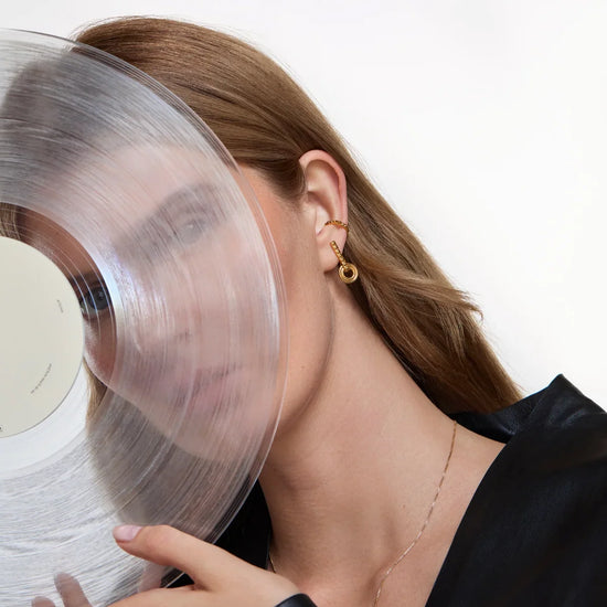 A close-up lifestyle shot of a woman with long light brown hair wearing a gold hoop earring and holding a translucent vinyl record against her face, wearing a black satin top against a clean white background.