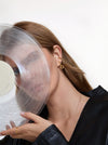 A close-up lifestyle shot of a woman with long light brown hair wearing a gold hoop earring and holding a translucent vinyl record against her face, wearing a black satin top against a clean white background.
