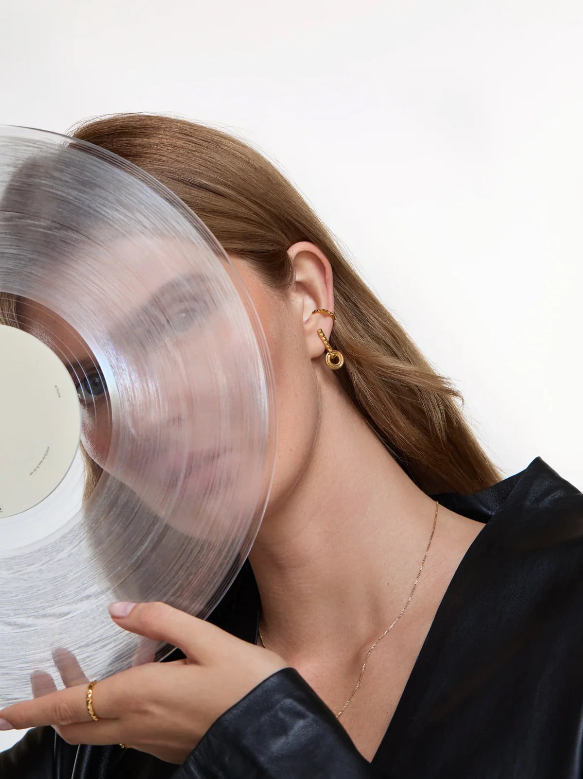 A close-up lifestyle shot of a woman with long light brown hair wearing a gold hoop earring and holding a translucent vinyl record against her face, wearing a black satin top against a clean white background.