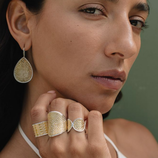Woman wearing gold earrings and ring against a neutral background from Anna Beck